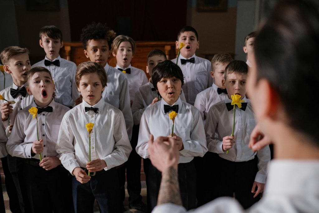 A boys' choir practicing together, each holding a yellow flower, led by a conductor over the shoulder.